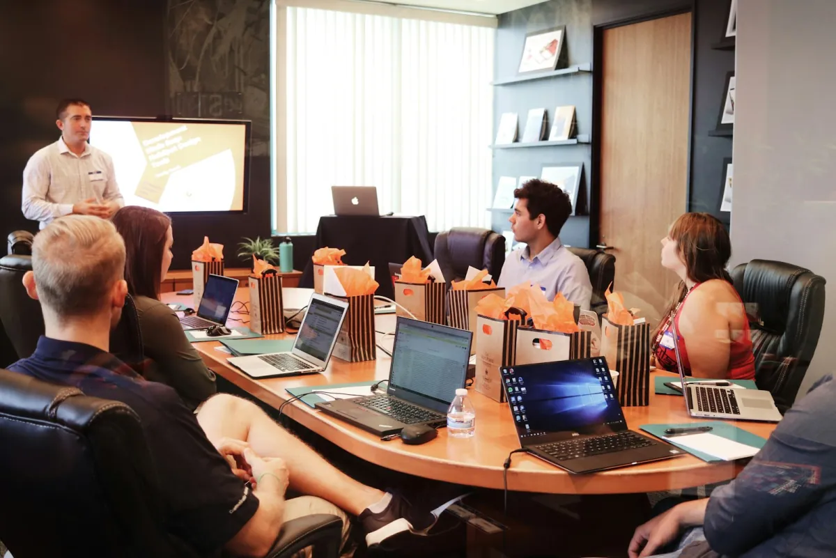 A presenter standing before a seated audience with open laptops in a meeting room, a slide on the screen behind them — an in-person interactive presentation