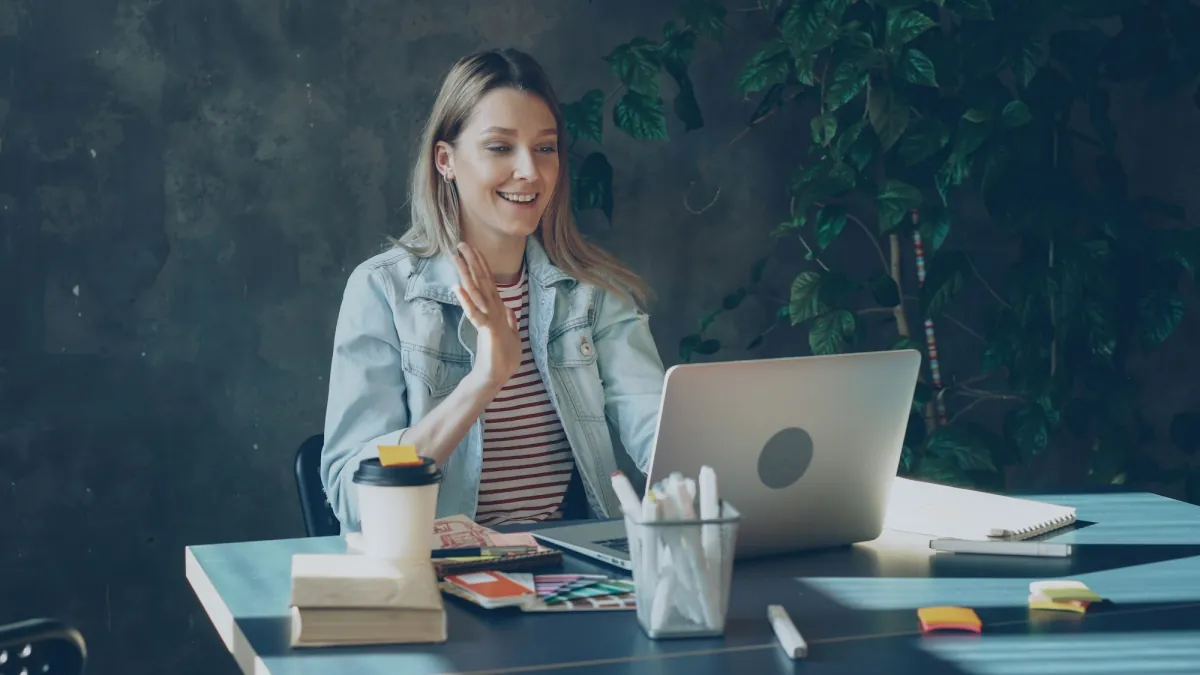 A remote worker smiling and waving at her laptop during a video call from home, ready to join a virtual pub quiz